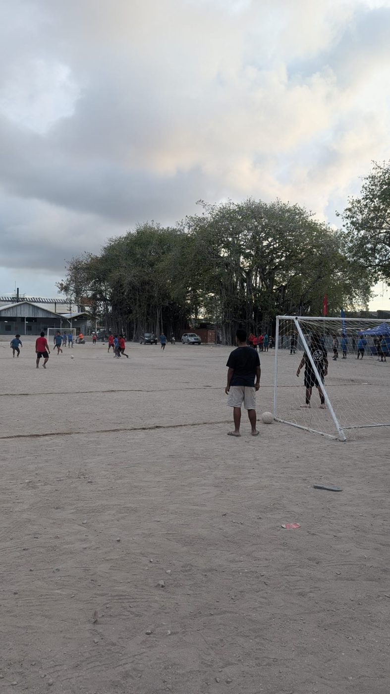 Children playing football towards a small goal on a sandy field on Naoero